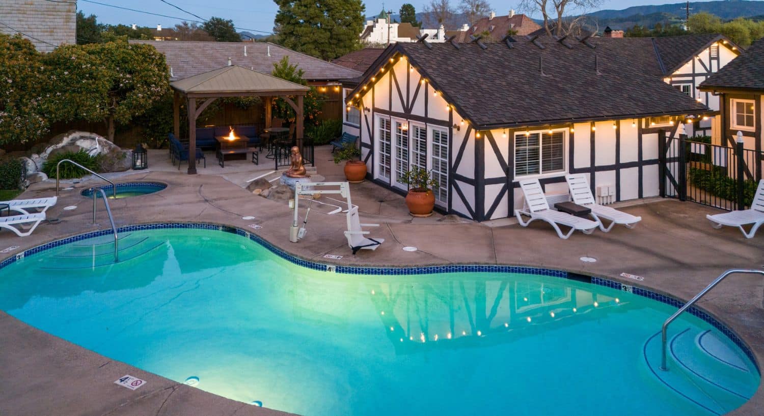 A curved outdoor pool area with lounging chairs, outdoor seating and firepit table and brown and white building
