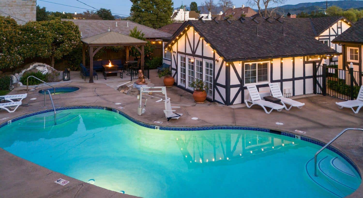 A curved outdoor pool area with lounging chairs, outdoor seating and firepit table and brown and white building