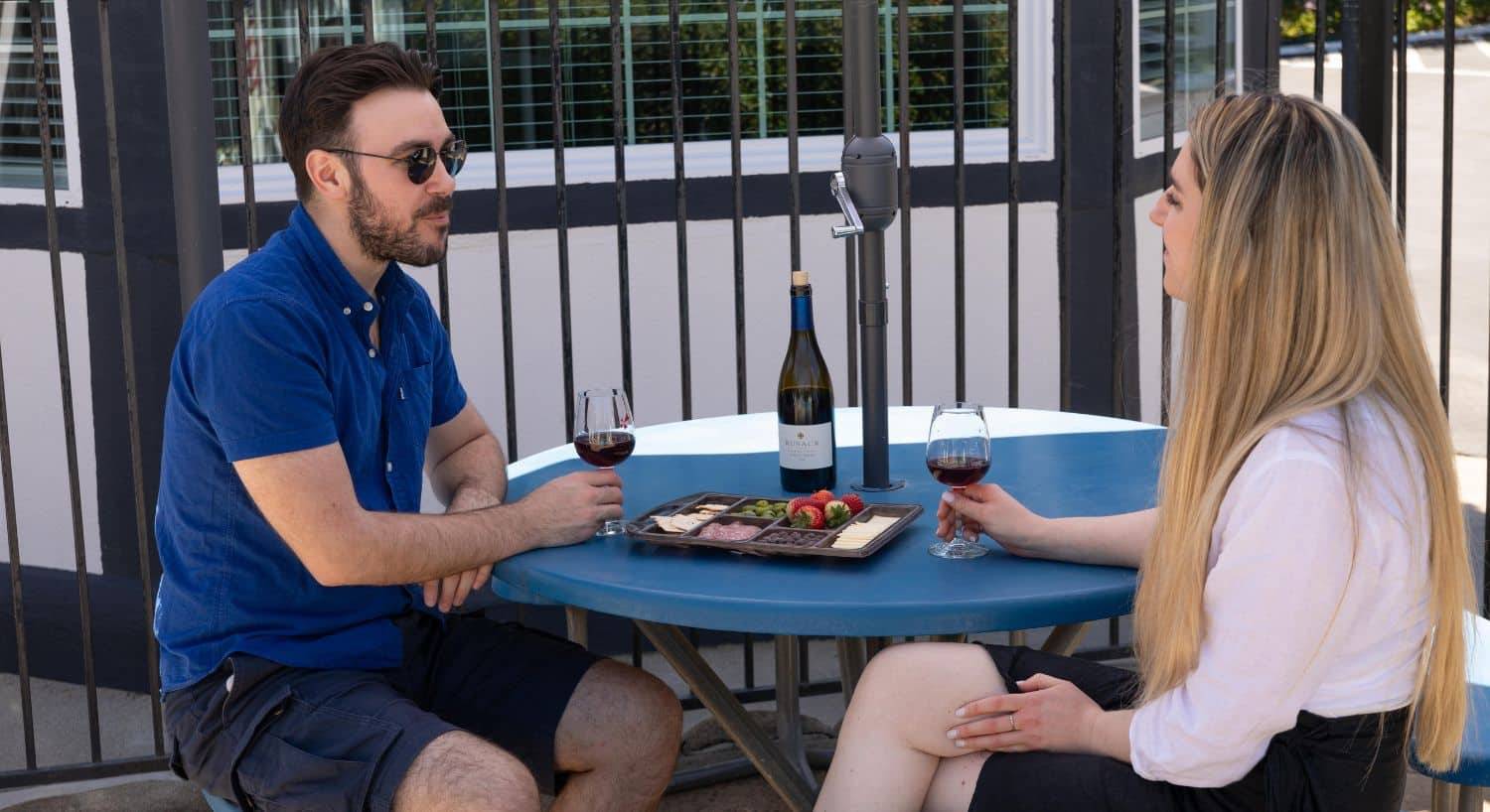 A man in a blue shirt with a woman in a white blouse sitting at an outdoor patio table enjoying wine and charcuterie