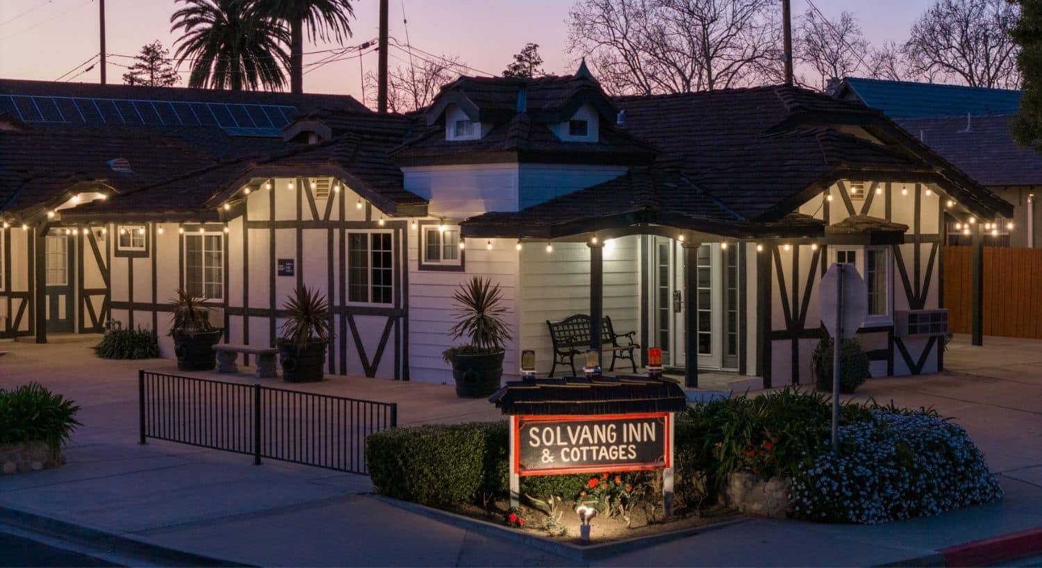 A large white building with brown trim lit up at night with white lights, red and black business sign out front with green bushes