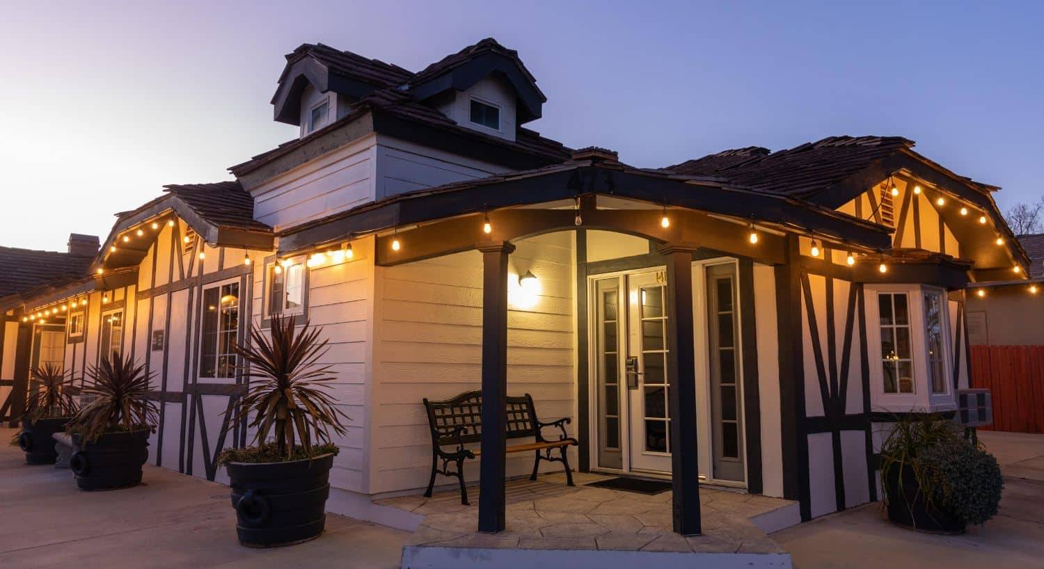 A large white building with brown trim lit up at night with white lights, bench by the front door and several potted plants,
