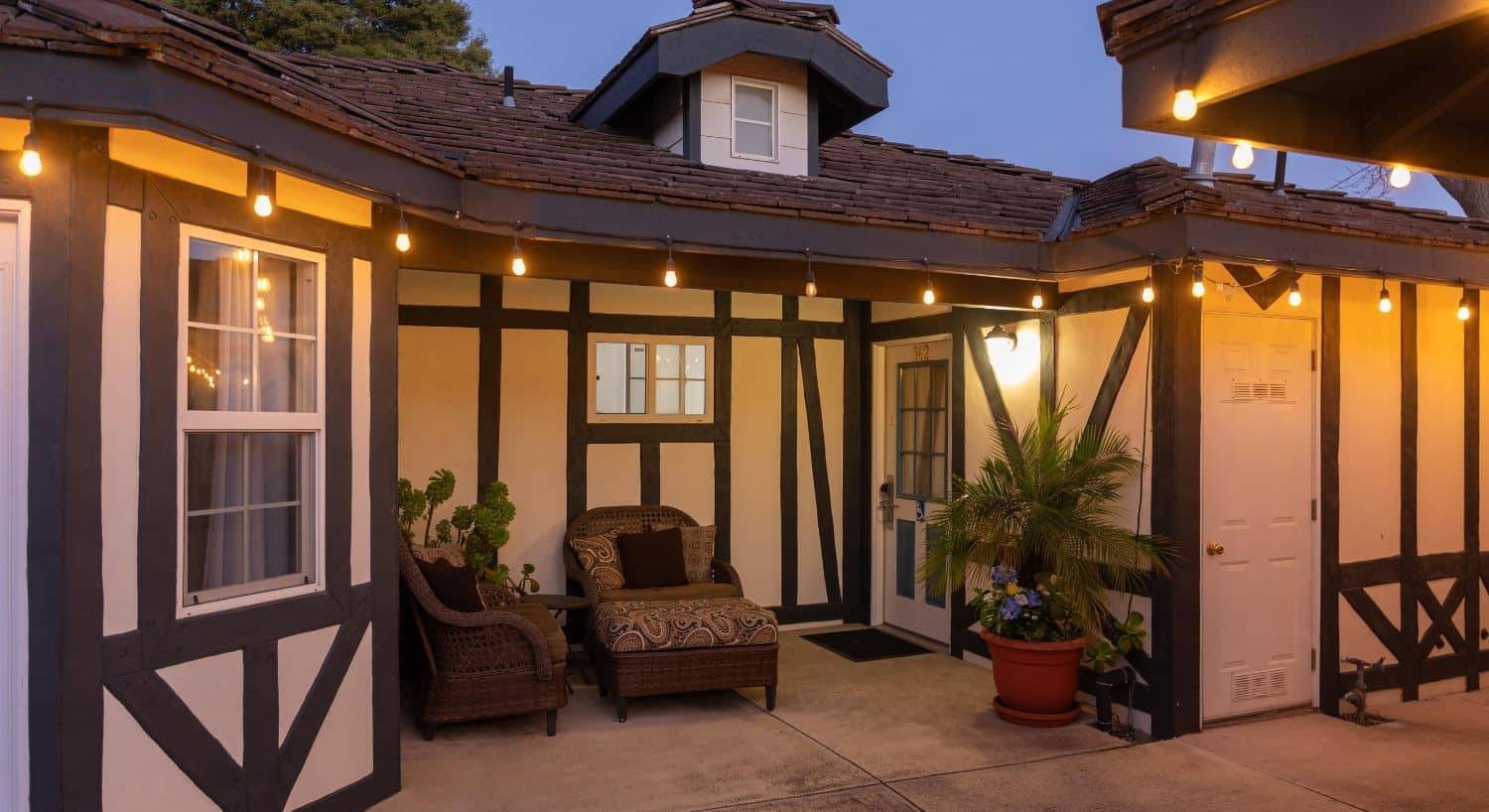 A white building with brown trim, outdoor seating by a front door and patio lights lit up at night