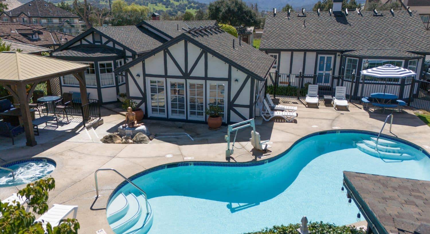 A large curved outdoor pool area nestled in between several white buildings with brown trim, with an outdoor patio area under a gazebo